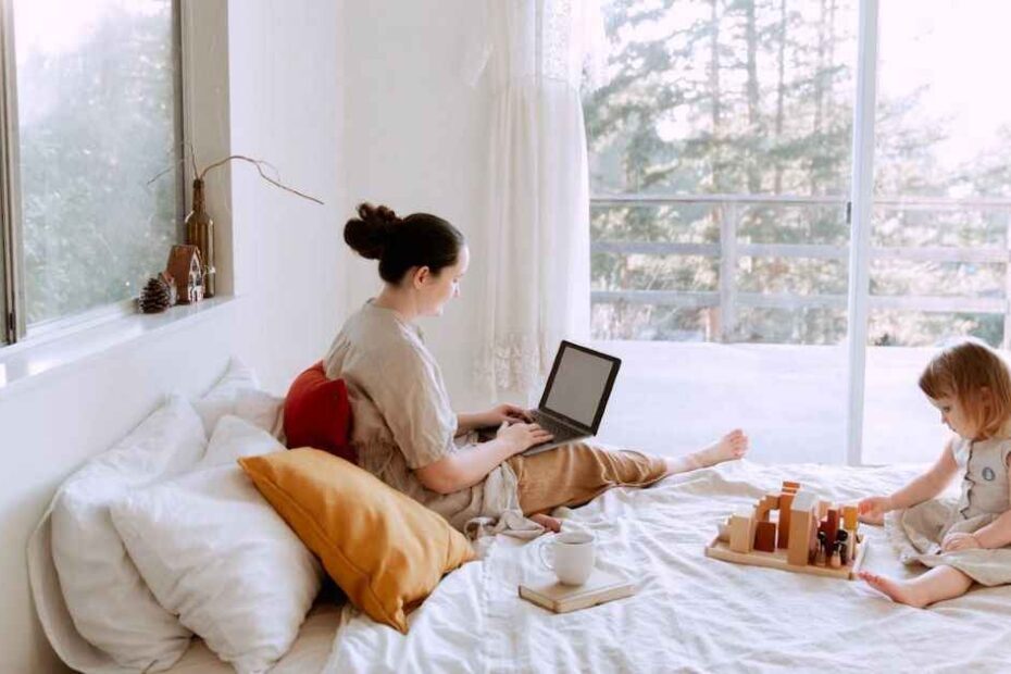 Adorable toddler girl playing with wooden blocks sitting on bed while mother using laptop on sunny morning